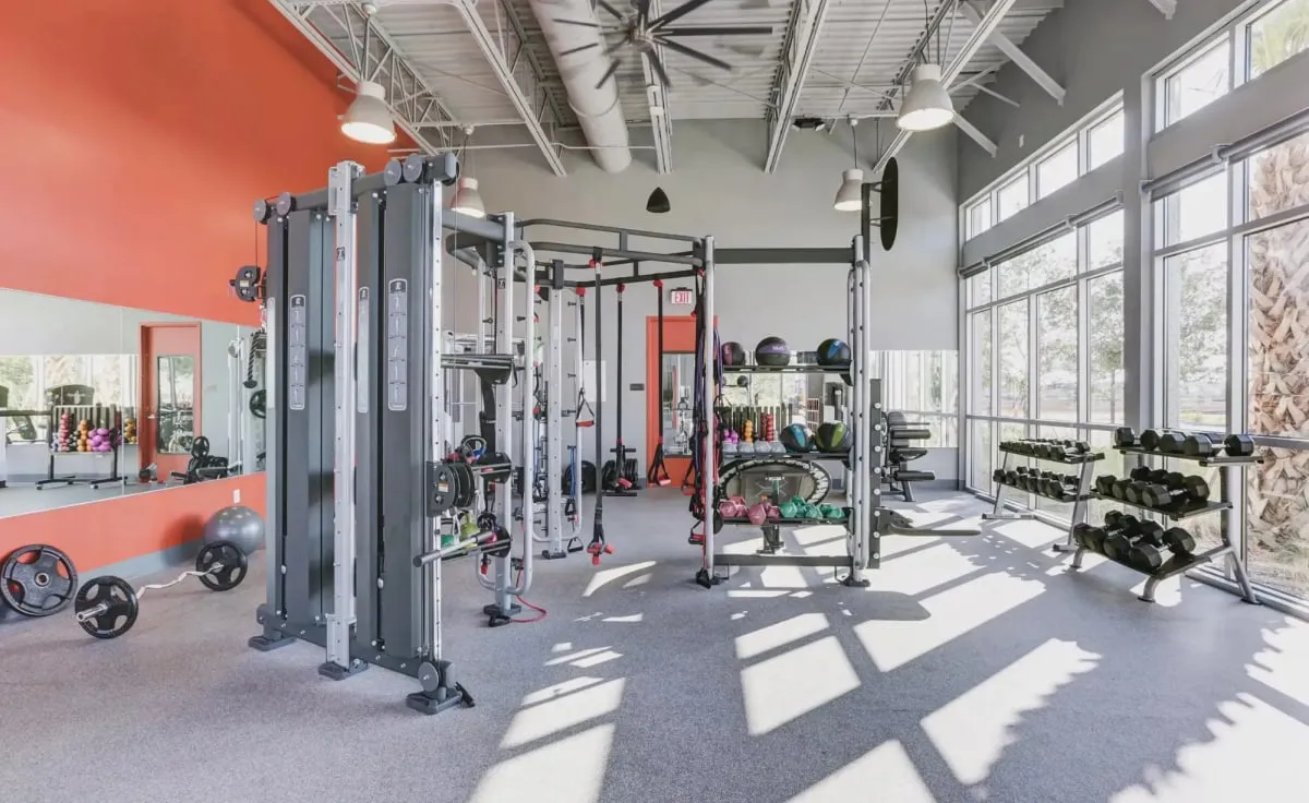 The Laureate Park fitness room: a cable machine and training rig, racks of dumbbells and medicine balls, an orange accent wall and floor-to-ceiling windows