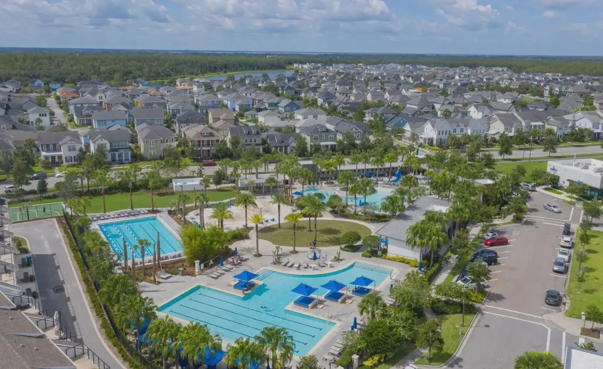 Aerial view of Laureate Park’s pools: two lap pools, a resort pool with blue cabana umbrellas and a splash area, a lawn with palm trees and a basketball court, with streets of homes and a lake beyond