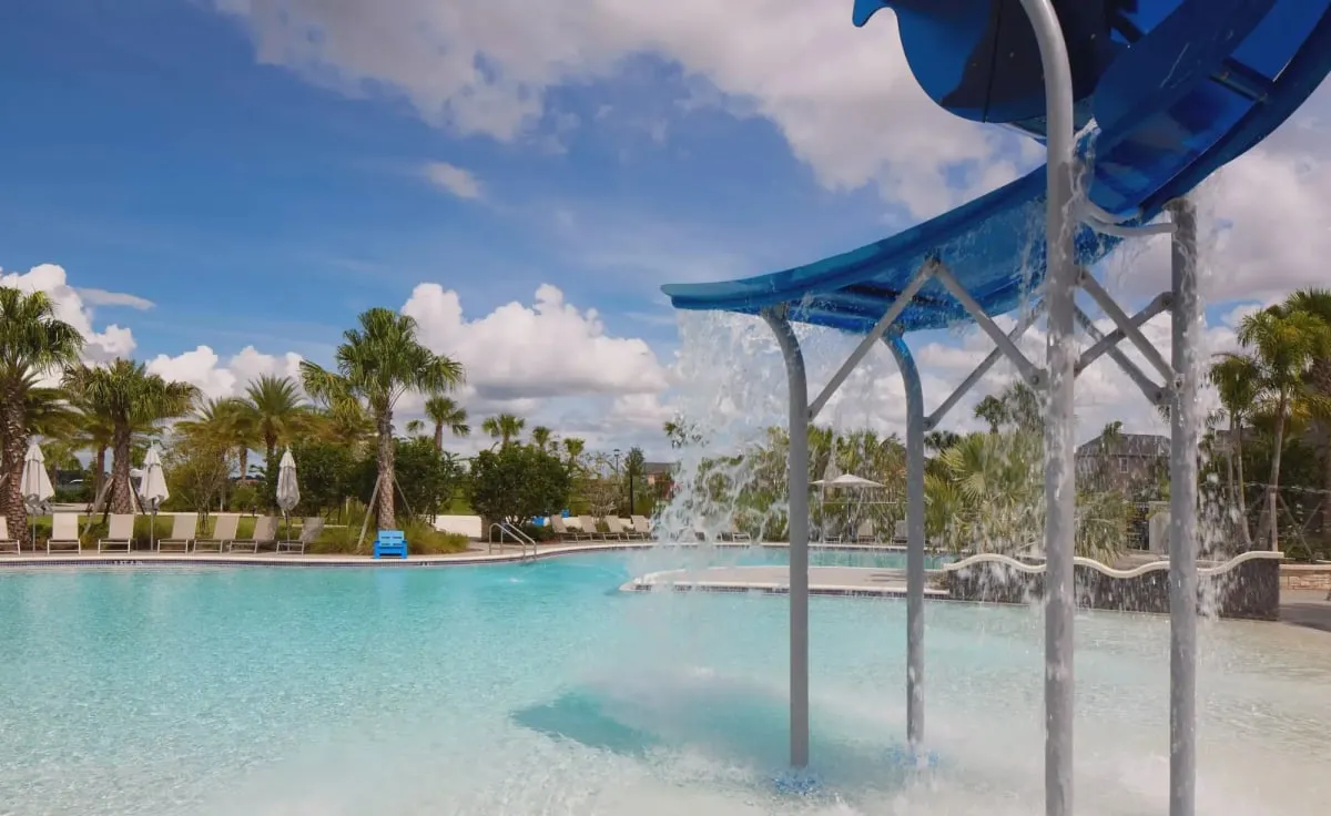 The Laureate Park splash pool: water pouring from a blue slide into a shallow beach-entry pool, with lounge chairs and palm trees behind
