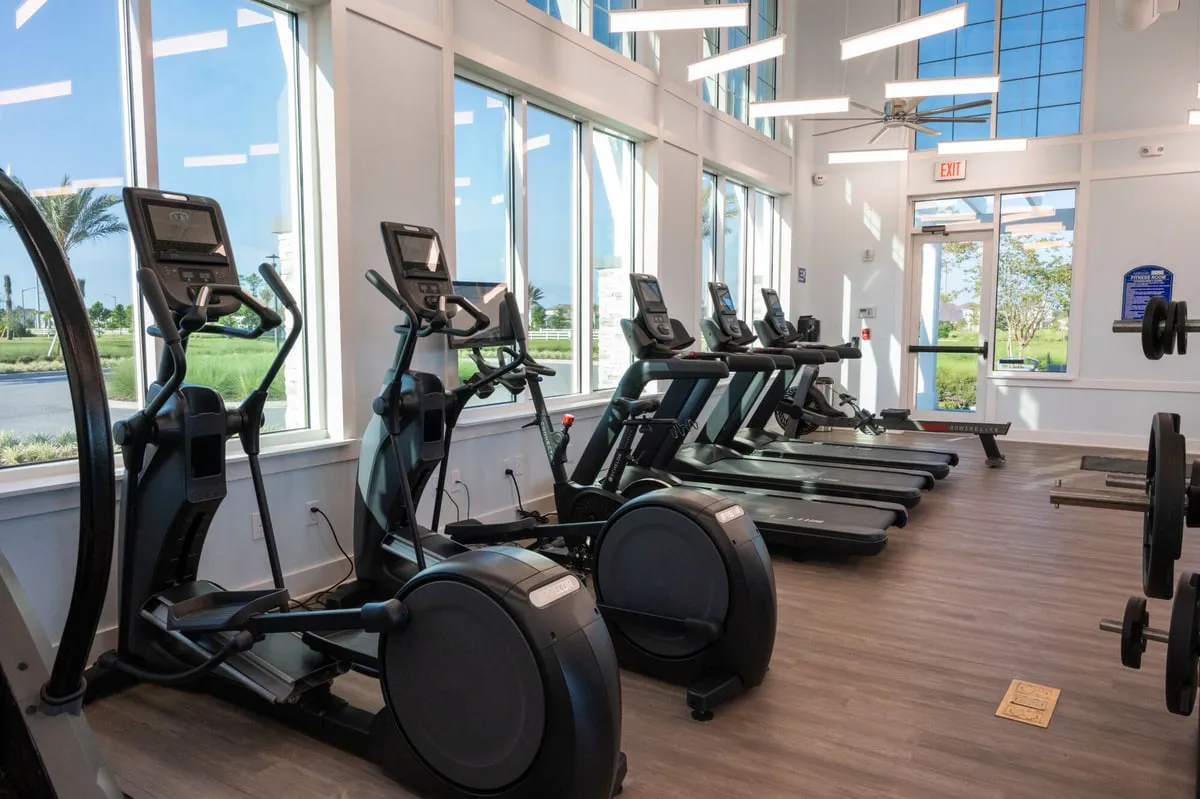 A row of ellipticals, an exercise bike, treadmills and a rowing machine along tall windows in the Meridian Parks fitness room, with palm trees and homes outside