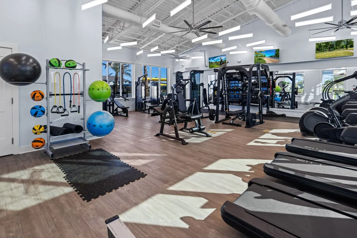 The Meridian Parks fitness room: treadmills, ellipticals, weight machines and a rack of exercise balls and resistance bands under a high white ceiling with fans, with windows onto the pool