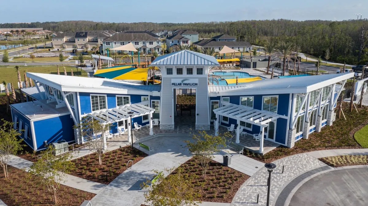 Aerial view of a blue amenity building with white trim and a central gate tower signed Pillar Pavilion, with shaded play areas, the pool and rows of homes behind it