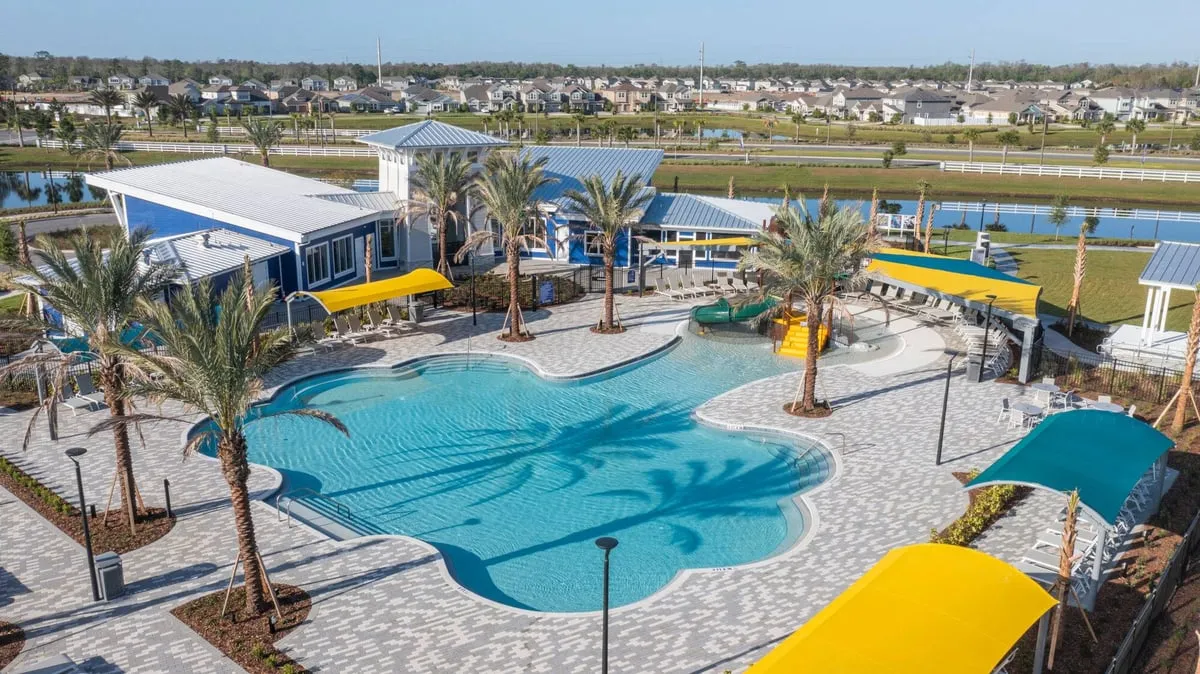 Aerial view of the Meridian Parks pool, with a children’s water slide, yellow and teal shade canopies and palm trees, next to a blue amenity building, with a lake and rows of homes behind it